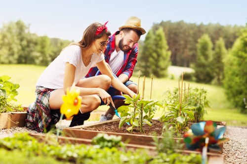 In the image, a young woman and a man, likely in a front or back garden of a residential property in Muswell Hill, are engaged in gardening activities. They are crouched down near a raised garden bed made of timber, which contains young, leafy green plants growing in rich soil. The woman, wearing a white t-shirt, plaid shorts, and a pink headband, is actively tending to the plants, while the man, dressed in a casual plaid shirt and a straw hat, is assisting or observing. The garden area includes a well-maintained lawn with short, dense grass visible in the foreground, and a gravel pathway surrounding the raised bed. In the background, there are tall, lush trees and a bright, sunny sky indicating clear weather. Decorative garden elements, such as small ornamental figures or plant markers, are also visible in the scene. The overall setting appears to be a spacious, well-kept outdoor space suitable for gardening and outdoor leisure, typical of residential gardens in the Muswell Hill area, where outdoor maintenance and horticultural services are often sought by homeowners. This scene reflects a peaceful gardening moment, aligning with services offered by Hedge Trimming Muswell Hill for garden tidiness and plant care.
