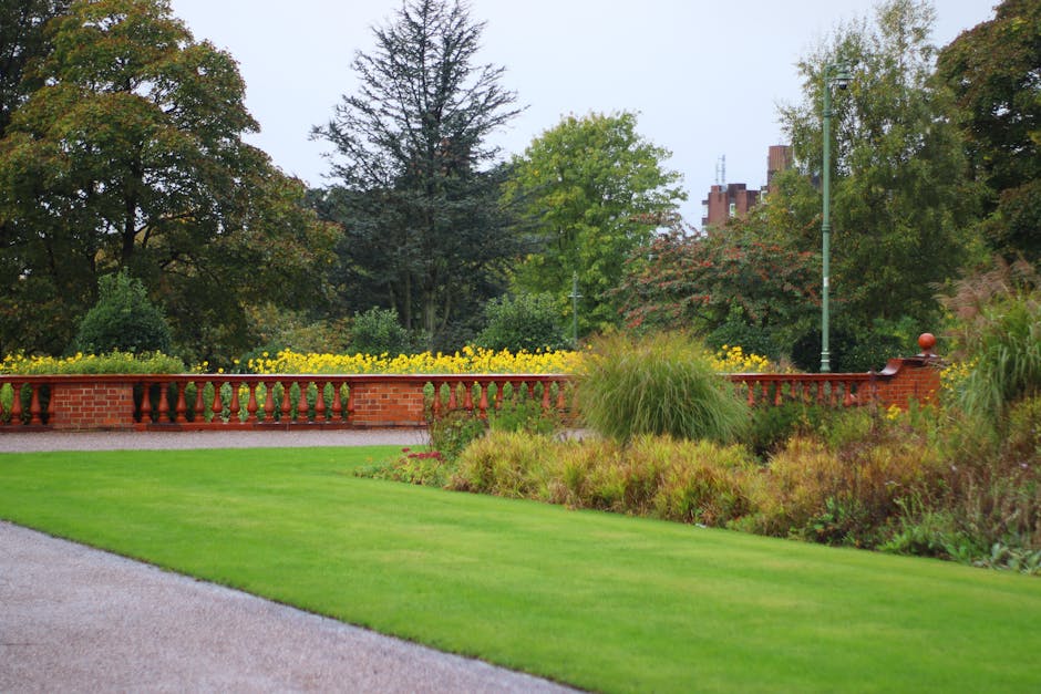 A large glass conservatory with curved, multi-pane panels and a rounded central dome, set in a well-maintained garden under a clear blue sky. The garden features neatly trimmed grass lawns with define