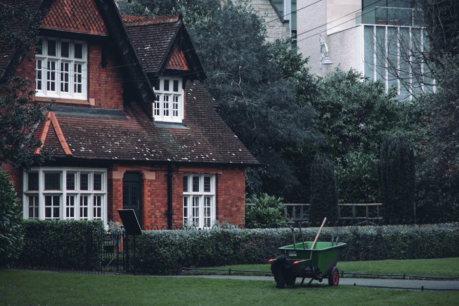 Two gardening workers in green jackets are engaged in outdoor maintenance in a park setting. One worker is pushing a blue wheelbarrow filled with dark soil or compost along a paved pathway, while the